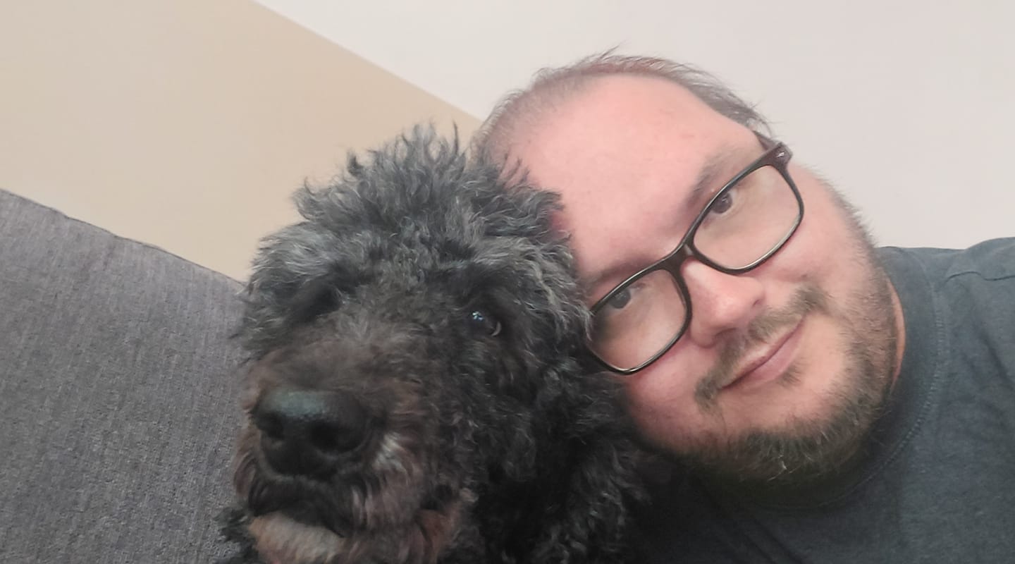 A man sits next to a black standard poodle, she is loooking at the camera